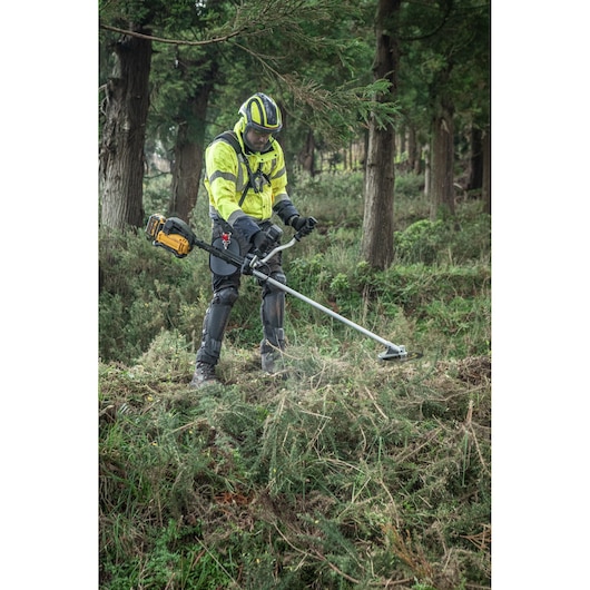 Operator uses DEWALT DCMBC723N-XJ, DCMBC723N brush cutter in forest. Safety gear worn.