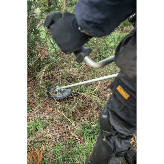DEWALT brush cutter in use outdoors, SKUs DCMBC723N-XJ, DCMBC723N. Grass and bushes visible.