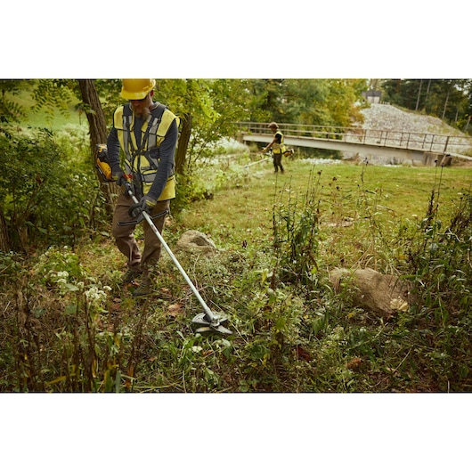 Two people using DEWALT DCMBC812N-XJ and DCMBC812N brush cutters outdoors, trimming grass near trees and a bridge.