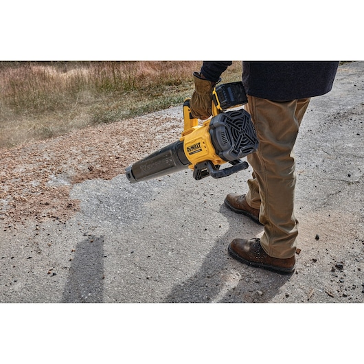 Person using a DEWALT cordless blower to clear debris from a paved outdoor surface.