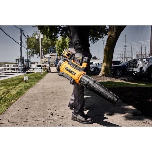 Person using a yellow DEWALT cordless leaf blower outdoors on a sidewalk.