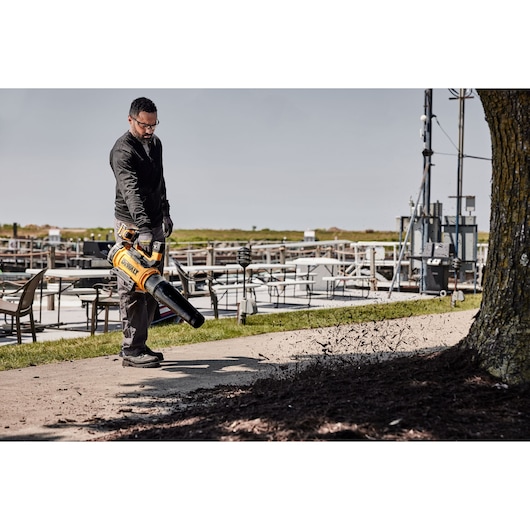 Person uses a DEWALT cordless leaf blower outdoors to clear debris from a sidewalk near tables and a tree.