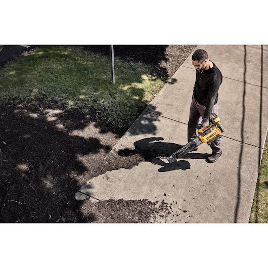 Person uses a DEWALT cordless leaf blower to clear debris from a sidewalk near grass and mulch.
