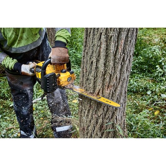 A person using a yellow DEWALT chainsaw to cut down a tree, with wood chips flying, in an outdoor forested area.