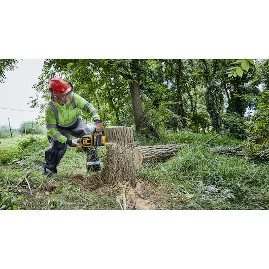 A person wearing safety gear uses a DEWALT cordless chainsaw to cut a fallen tree trunk in a forested outdoor area.
