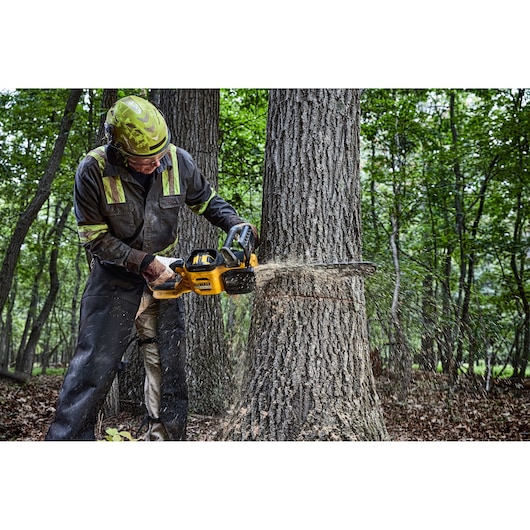 A person wearing protective gear is cutting a tree trunk in a forest using a yellow DEWALT chainsaw, model DCMCS575N. Wood
