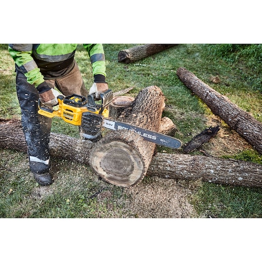 A person using a DEWALT cordless chainsaw to cut a large log outdoors, with wood chips and sawdust visible around the area.
