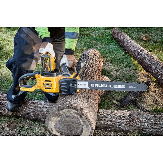 A person using a DEWALT 54V XR FLEXVOLT brushless cordless chainsaw to cut a large log outdoors. The chainsaw is yellow and black, and the person is wearing gloves and protective clothing.