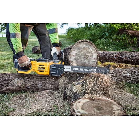 A person using a yellow DEWALT cordless brushless chainsaw to cut through a large tree log outdoors. Sawdust is visible flying as the chainsaw operates.