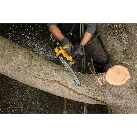 Person uses a DEWALT chainsaw to cut a thick tree branch outdoors, wearing gloves and safety gear.