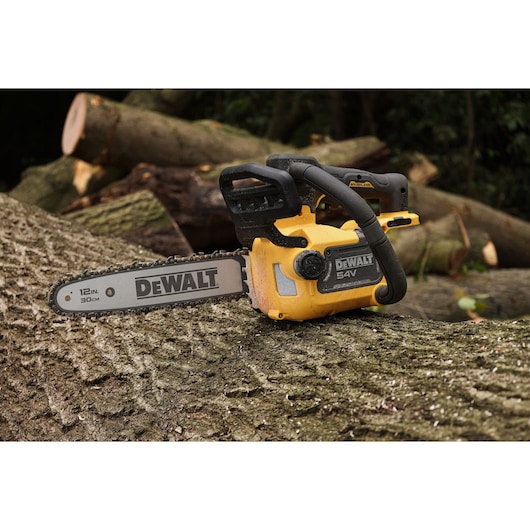 A DEWALT 54V cordless chainsaw with a 12-inch (30 cm) bar, shown resting on a large tree trunk outdoors, with cut logs in the background.