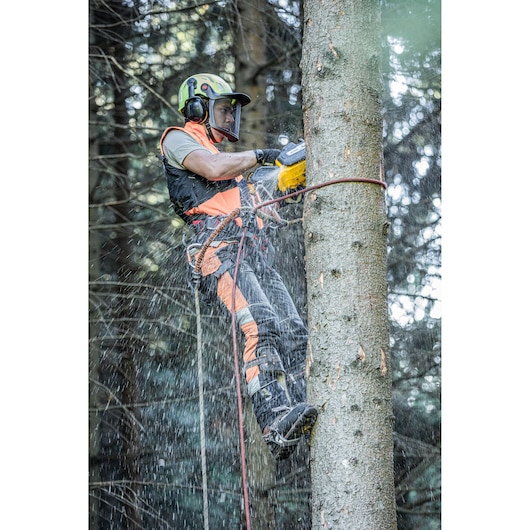 Worker uses a DEWALT chainsaw to cut a tree while climbing with safety gear in a forest, wood chips flying.