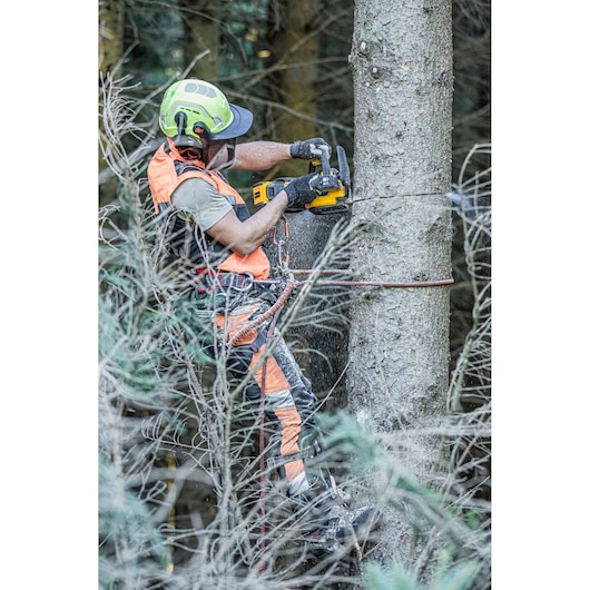 Worker in safety gear uses a DEWALT chainsaw to cut a tree trunk in a forest.