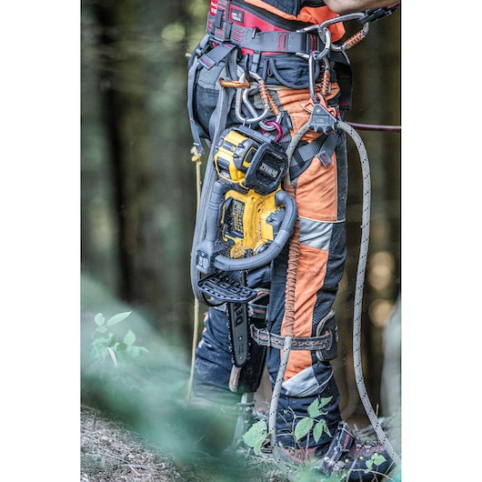 A DEWALT chainsaw attached to a worker's harness and belt, shown outdoors with safety gear.