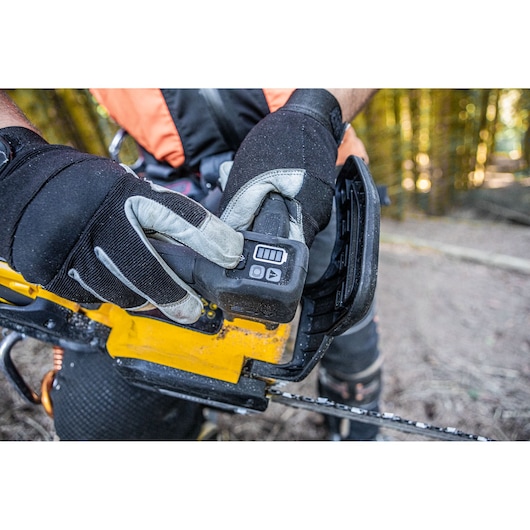 Gloved hands inserting the battery into a yellow DEWALT chainsaw outdoors on a forest floor.