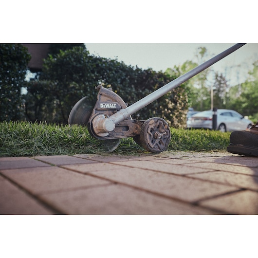 DEWALT lawn edger trims grass along a sidewalk with bushes and parked cars in the background.