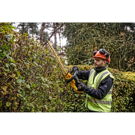 Person trims hedge outdoors with a yellow DEWALT cordless hedge trimmer, wearing protective gear and safety vest.