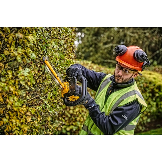 Person trims hedge with DEWALT cordless hedge trimmer, wearing safety gear and high-visibility vest outdoors.