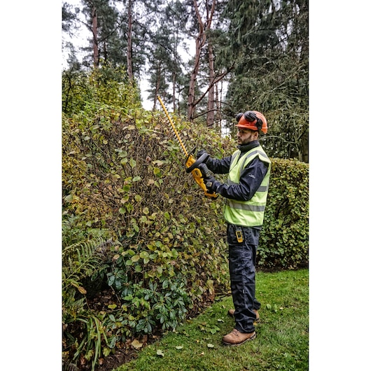 Worker trims hedge outdoors using DEWALT DCMHT563P1 cordless hedge trimmer; wearing safety vest and helmet.
