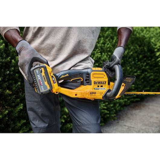 A person wearing gloves is holding a DEWALT 54V cordless hedge trimmer outdoors, with green bushes in the background.