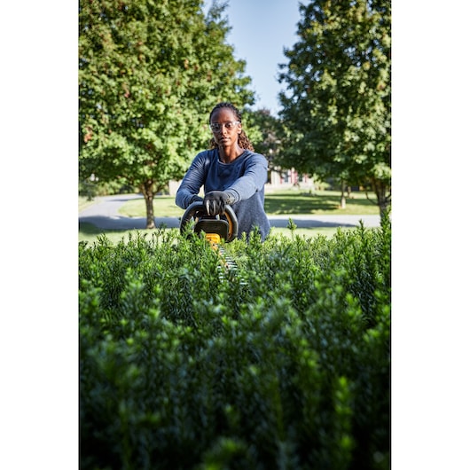 Person using a DEWALT hedge trimmer to cut bushes outdoors, surrounded by greenery and trees.