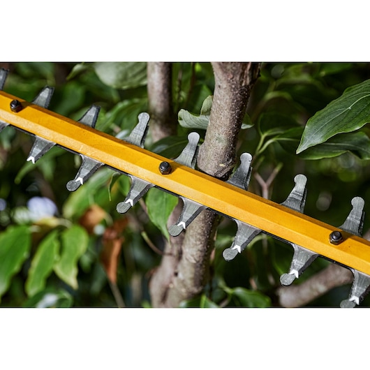 Close-up of DEWALT hedge trimmer blades cutting a tree branch among green leaves.