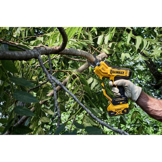Person uses DEWALT cordless pruner to cut a tree branch outdoors amid green leaves.