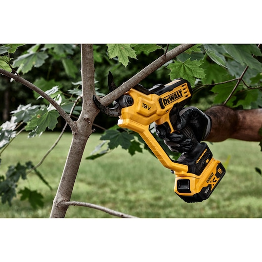 A DEWALT 18V cordless pruner is cutting a tree branch outdoors, held by a gloved hand.