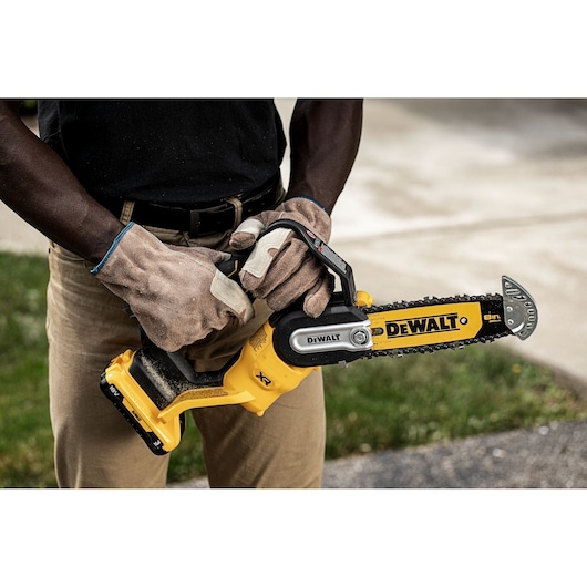 Person wearing gloves holding a DEWALT cordless chainsaw outdoors close to their body.