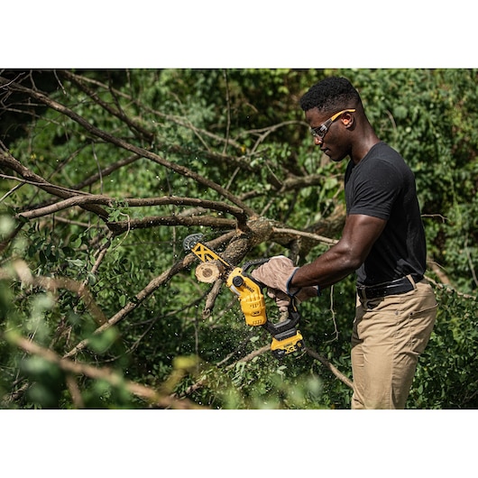 Person uses a DEWALT cordless chainsaw to cut tree branches outdoors. Face blurred for privacy.