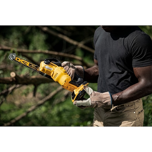 Person wearing gloves holds a DEWALT cordless chainsaw outdoors, with trees in the background.