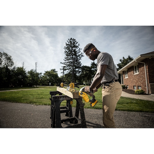 Person sawing wood outdoors with a DEWALT cordless chainsaw on a workbench near a brick house.