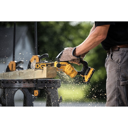Person using a DEWALT cordless chainsaw to cut wood outdoors, sawdust flying around.