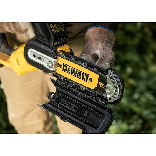 Close-up of a DEWALT cordless chainsaw held by gloved hands, with the chain guard open outdoors.