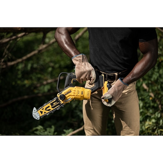 Person wearing gloves holds a DEWALT cordless chainsaw outdoors, preparing to use it near greenery.