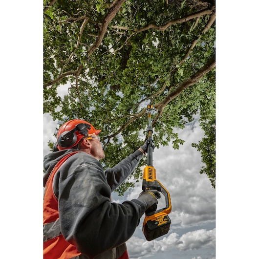 Person uses a DEWALT cordless pole saw to trim tree branches outdoors, wearing safety gear under cloudy sky.