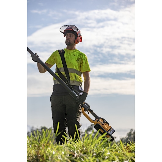 Worker outdoors holding a DEWALT DCMPS640N pole saw, wearing safety gear and yellow shirt under a bright sky.