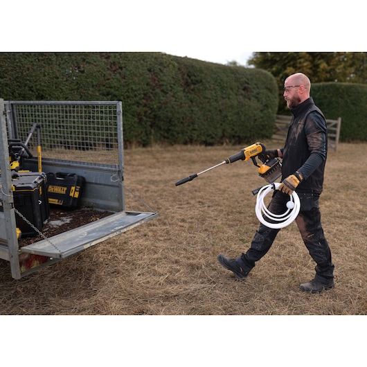 Man uses DEWALT DCMPW1000N, DCMPW1000N-XJ pressure washer outdoors, holding hose and tool near trailer.