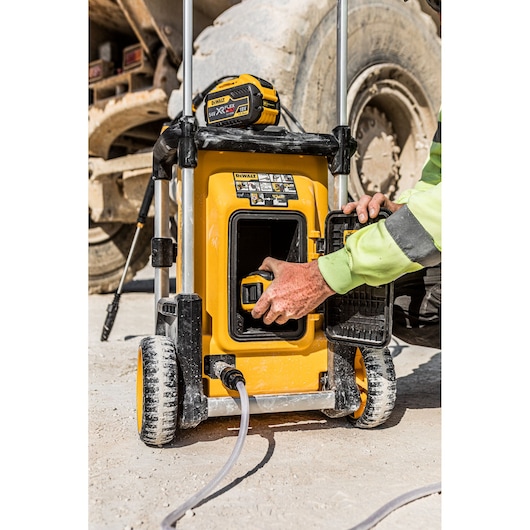 Person inserts battery into yellow DEWALT DCMPW1600 portable pressure washer near construction equipment.