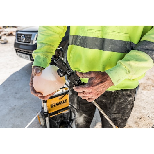 Person attaches soap bottle to DEWALT pressure washer, wearing a high-visibility jacket at a worksite.
