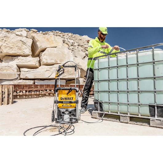 DEWALT DCMPW1600 pressure washer connected to water tank outdoors, worker in high-visibility gear nearby.