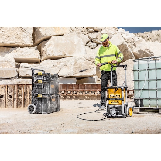 DEWALT DCMPW1600 pressure washer cleaning toolboxes outdoors near rocks and water tank.
