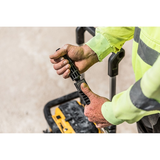 Close-up of hands adjusting a DEWALT tool handle outdoors with a yellow power tool in the background.
