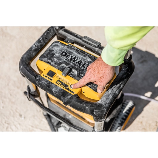 Hand presses button on DEWALT portable pressure washer DCMPW1600 at a dusty outdoor site.