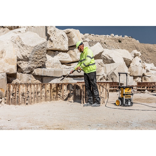 Worker uses DEWALT pressure washer outdoors near large rocks and metal structure.