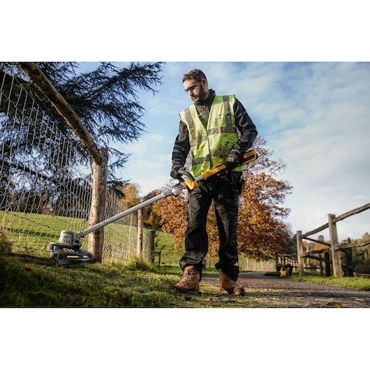 Person uses DEWALT DCMST561N grass trimmer outdoors near a fence on a sunny day.
