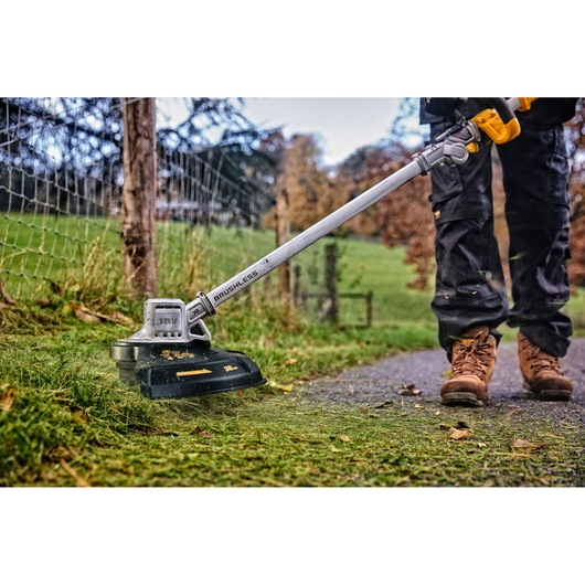 DEWALT cordless brushless grass trimmer used outdoors near a fence, operator in work boots and pants visible.