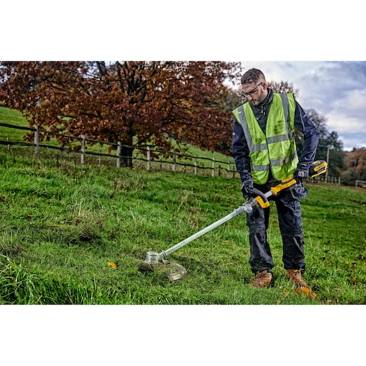 Person uses a DEWALT cordless grass trimmer outdoors, wearing a safety vest on green grassy terrain.