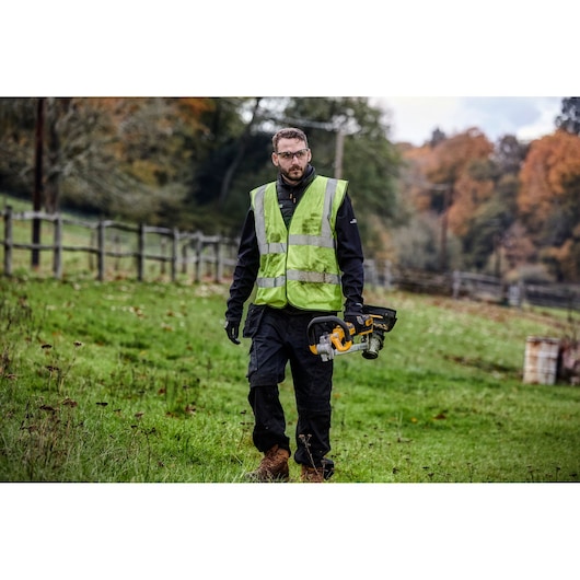 Person outdoors holding a DEWALT grass trimmer, wearing a safety vest and work boots in a grassy area.