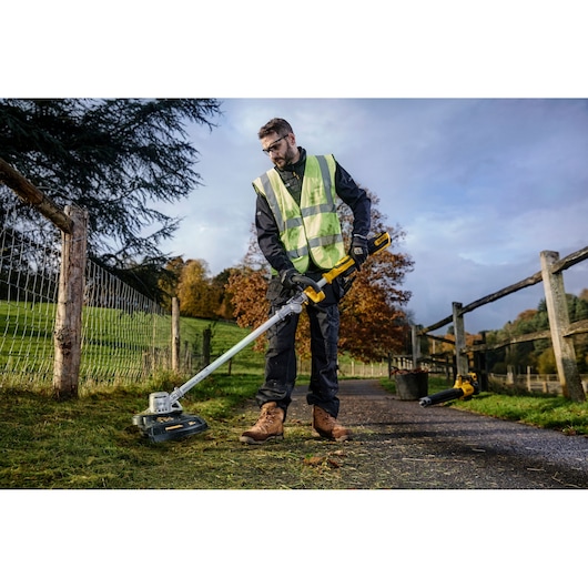 Person using a DEWALT cordless grass trimmer outdoors, wearing safety gear and working beside a wooden fence.